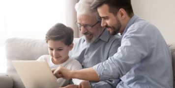 Happy grandfather and father teaching little boy to use laptop, sitting together on couch, three generations of men playing game, shopping or chatting online, having fun with gadget at home